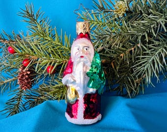 May include: A vintage Christmas ornament shaped like Father Christmas, with a white beard and red suit. He holds a green tree and a gold sack. The ornament is decorated with white glitter and a gold star on top. The background includes pine needles, red berries, and a pine cone.