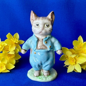 May include: A ceramic cat figurine wearing a light blue jacket and matching trousers, standing on a small green base. The cat has white and brown fur, pointed ears, and is positioned between two clusters of bright yellow daffodil flowers.