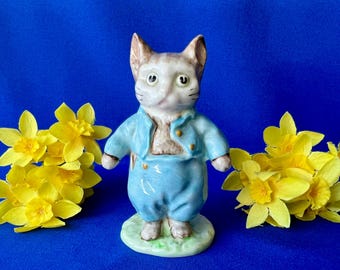 May include: A ceramic cat figurine wearing a light blue jacket and matching trousers, standing on a small green base. The cat has white and brown fur, pointed ears, and is positioned between two clusters of bright yellow daffodil flowers.