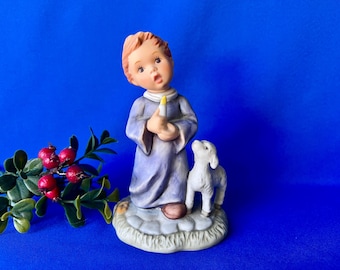 May include: A ceramic figurine of a child with light brown hair wearing a long, light purple robe, holding a small white candle with a yellow flame. A small white lamb stands beside the child on a textured, stone-like base.