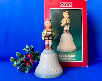 May include: A ceramic bell featuring a figurine of a boy wearing a green jacket, brown shorts, and a red scarf. The bell is light grey with embossed flower details. It is displayed with its original red box labelled Schmid Hark the Herald.