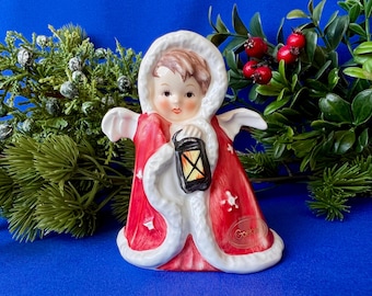 May include: A ceramic figurine of a child wearing a red hooded cloak with white trim, holding a black lantern. The cloak features white star accents and a gold Goebel stamp. The child has brown hair and is depicted with wings.