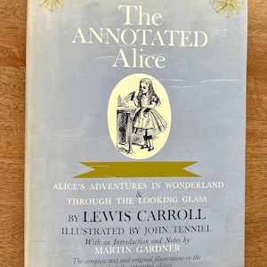 May include: A vintage hardcover book titled "The Annotated Alice." The cover is light blue with gold accents and features an illustration of Alice. Text includes the author, illustrator, and the book's full title: "Alice's Adventures in Wonderland & Through the Looking Glass."