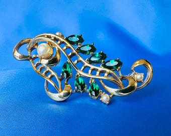 May include: A gold-toned brooch with a delicate, intricate design featuring a leafy motif. The brooch is adorned with emerald green and white faux pearls.