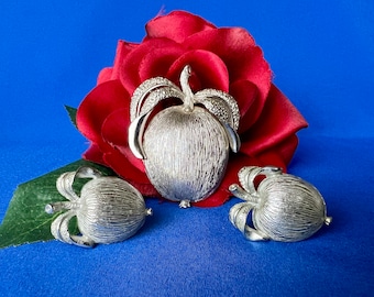 May include: A silver tone brooch and earrings set featuring a textured, stylised fruit design. The brooch is larger than the earrings and has a more elaborate design. The earrings are simple and elegant.