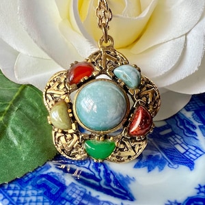 May include: A vintage gold-tone pendant necklace with a central light blue stone surrounded by smaller red, green, and blue-green stones. The pendant has an ornate, openwork design and hangs on a delicate gold chain. The necklace is displayed on a white flower and blue and white patterned surface.