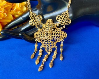 May include: A gold-tone necklace featuring a central filigree pendant with an intricate lattice pattern. The pendant is adorned with five dangling, textured drop-shaped charms suspended from the base, connected to a delicate gold-tone chain.