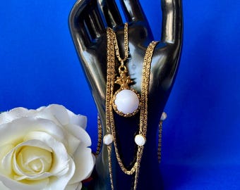 May include: A gold-tone multi-strand necklace featuring a central pendant with a round white cabochon stone and smaller matching white stone accents along the chain links. The necklace has a textured metal finish and a decorative crown-like setting.