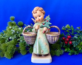 May include: A ceramic figurine of an angel with light brown hair, wearing a pale green robe decorated with small stars. The angel holds a small green Christmas tree in one hand and carries two brown woven baskets on its arms.