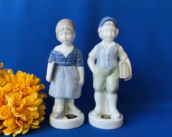 May include: Two porcelain figurines of children standing on circular bases. The left figure wears a blue dress and pink cap, holding a book. The right figure wears a blue vest, green shorts, and blue cap, holding a book. Both have white skin.