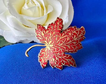May include: A red and gold maple leaf brooch with detailed patterns. The brooch is set against a blue background with a white rose in the upper left corner. The leaf design features a gold filigree pattern.