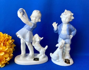 May include: Two white porcelain figurines featuring a child in a blue dress holding a ball with a dog, and a child in a blue outfit with a dog. Both figures stand on oval bases with black and gold labels featuring a crown and the letters ELR.