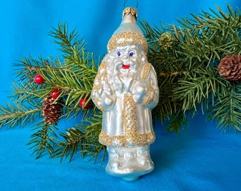 May include: A vintage Christmas ornament in the shape of Father Christmas. The silver ornament has gold glitter detailing on the hat, coat trim, and cuffs. Father Christmas has blue eyes and a red mouth. It is displayed with pine branches and a blue background.
