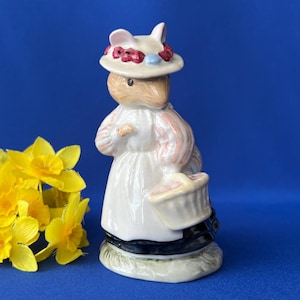 May include: A ceramic figurine of a mouse wearing a hat and carrying a basket, with yellow daffodils on the left. The mouse is dressed in a white dress with pink and blue accents. The figurine is on a blue background.