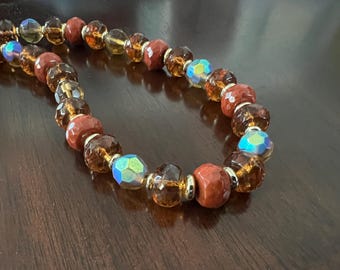 May include: A beaded necklace featuring a repeating pattern of faceted amber-coloured glass beads, opaque terracotta-coloured round beads, and iridescent blue-tinted faceted beads, all separated by small metallic gold-toned spacer rings.
