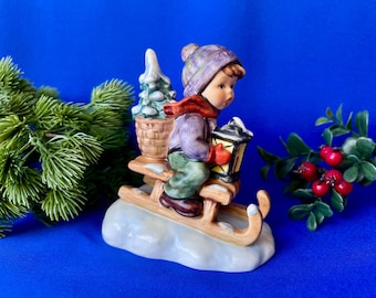 May include: A ceramic figurine of a child wearing a purple knit hat, purple jacket, and green trousers, riding a wooden sled over a snow-covered base. The child holds a lantern and carries a small basket containing a snow-dusted evergreen tree.