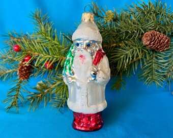 May include: A vintage Christmas ornament shaped like Father Christmas, with a white coat, red base, and a green tree. The ornament is set against a blue background with pine branches and a pine cone.