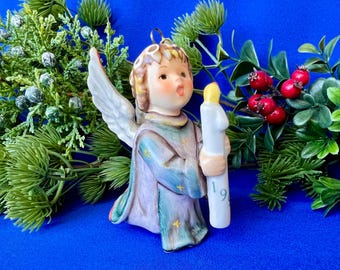 May include: A ceramic angel figurine with light brown hair and white wings, wearing a blue and purple robe decorated with small yellow stars. The angel holds a white candle with a yellow flame, featuring the number 1994 printed in green.