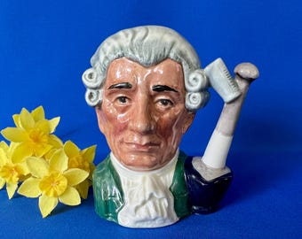 May include: A ceramic mug in the shape of a man's head, with a white wig and a green jacket. The mug has a gavel-shaped handle. Yellow daffodils are to the left of the mug against a blue backdrop.