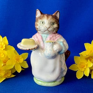 May include: A ceramic cat figurine wearing a pink shawl and white apron, holding a small plate with a yellow butter pat and a blue patterned pitcher. The cat has brown and white fur, green eyes, and is positioned between two yellow flowers.