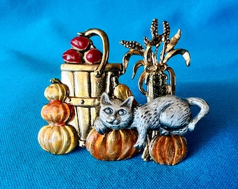 May include: A detailed brooch featuring a grey cat resting on a pumpkin, with a golden bucket of red berries and decorative corn stalks. The brooch has a fall theme with orange, gold, and red colours.