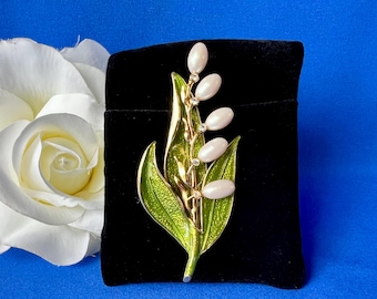 May include: A gold-tone metal brooch shaped like a lily of the valley plant. The piece features vibrant green enamelled leaves with a textured finish and five smooth, oval-shaped white faux pearls representing the flowers, accented by small clear crystals.
