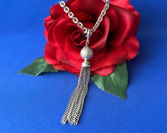 May include: A silver-toned necklace featuring a thick chain, a central spherical bead with a textured surface, and a long, multi-strand chain tassel pendant. The jewellery is displayed against a large, deep red fabric rose with green leaves.