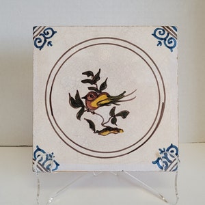 May include: Square ceramic tile with a hand-painted bird design. The bird is yellow, green, and brown, perched on a branch within a brown circle. The tile has blue corner accents and is displayed on a clear acrylic stand.