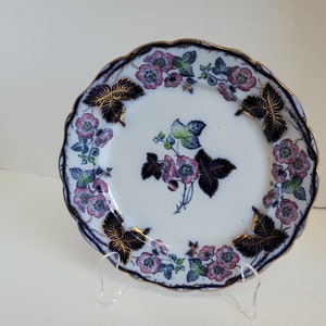 May include: A small white ceramic plate with a blue and pink floral design. The plate has a scalloped edge and a gold trim.