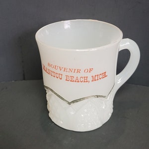 May include: White ceramic mug with a raised design around the bottom. The mug has a handle and says "Souvenir of Manitou Beach, Mich" in red lettering.