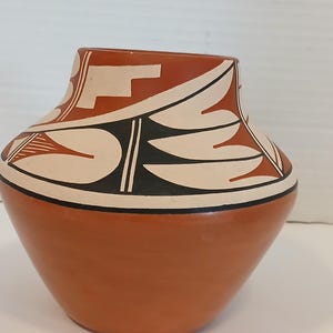 Puede incluir: Un jarrón de cerámica de color terracota con una base ancha y un cuello más estrecho. La parte superior presenta un diseño geométrico en crema, negro y marrón rojizo. El jarrón tiene un acabado liso y brillante.