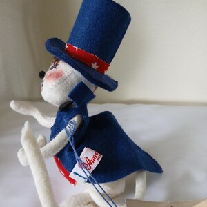Peut inclure: Poup&eacute;e lapin en feutre blanc faite &agrave; la main, portant un haut-de-forme bleu avec une bande rouge et blanche &eacute;toil&eacute;e, une cape bleue et une cravate rouge. La poup&eacute;e a des joues roses et une &eacute;tiquette "Anna Mer".