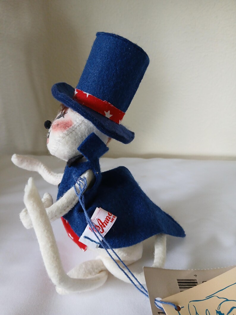 Peut inclure: Poup&eacute;e lapin en feutre blanc faite &agrave; la main, portant un haut-de-forme bleu avec une bande rouge et blanche &eacute;toil&eacute;e, une cape bleue et une cravate rouge. La poup&eacute;e a des joues roses et une &eacute;tiquette "Anna Mer".