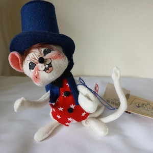 Peut inclure: Figurine de souris artisanale, v&ecirc;tue d'un haut-de-forme bleu, d'un gilet bleu marine et d'un gilet rouge orn&eacute; d'&eacute;toiles blanches. La souris a un visage peint avec des joues roses. Pos&eacute;e sur une surface blanche.