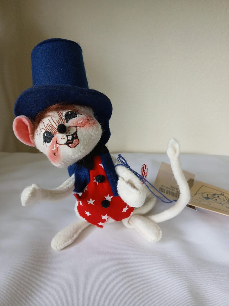 Peut inclure: Figurine de souris artisanale, v&ecirc;tue d'un haut-de-forme bleu, d'un gilet bleu marine et d'un gilet rouge orn&eacute; d'&eacute;toiles blanches. La souris a un visage peint avec des joues roses. Pos&eacute;e sur une surface blanche.