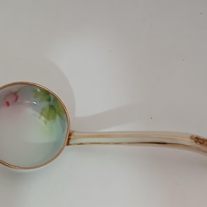 Puede incluir: Un cucharón de porcelana blanca con un borde y un mango dorados. El cuenco del cucharón está decorado con un diseño floral pintado a mano en tonos rosa, verde y azul.