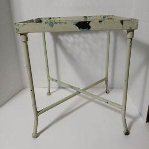 May include: A small, off-white metal plant stand with a rectangular top and four legs. The legs are connected by an X-shaped brace. The paint is distressed with areas of blue and black showing through.