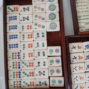 Puede incluir: Un juego de fichas de Mahjong de color marfil con varias marcas circulares y num&eacute;ricas de colores, dispuestas en bandejas de madera. Las fichas presentan c&iacute;rculos verdes, naranjas y azules, y caracteres chinos.