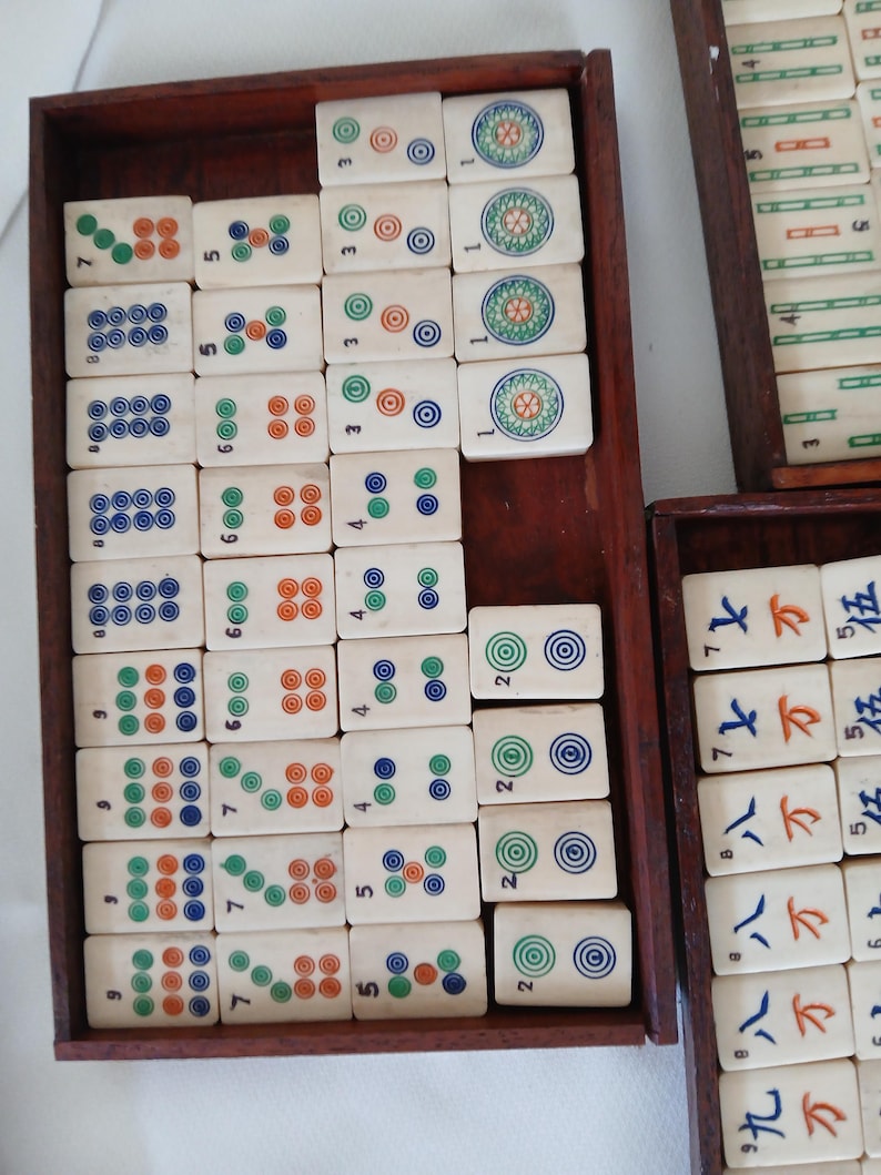 Puede incluir: Un juego de fichas de Mahjong de color marfil con varias marcas circulares y num&eacute;ricas de colores, dispuestas en bandejas de madera. Las fichas presentan c&iacute;rculos verdes, naranjas y azules, y caracteres chinos.