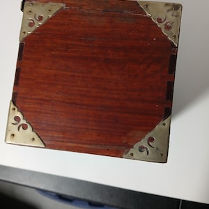 Puede incluir: Peque&ntilde;a caja rectangular de madera con un acabado marr&oacute;n rojizo intenso. Las esquinas est&aacute;n reforzadas con placas decorativas de metal color lat&oacute;n. La caja parece ser vintage o antigua, mostrando un dise&ntilde;o cl&aacute;sico.