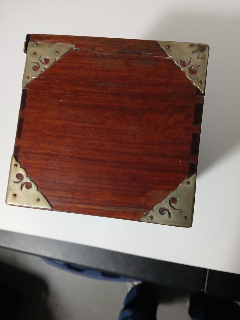 Puede incluir: Peque&ntilde;a caja rectangular de madera con un acabado marr&oacute;n rojizo intenso. Las esquinas est&aacute;n reforzadas con placas decorativas de metal color lat&oacute;n. La caja parece ser vintage o antigua, mostrando un dise&ntilde;o cl&aacute;sico.