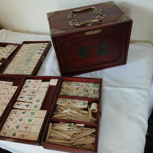 Puede incluir: Un juego de Mahjong de madera vintage con un acabado rojo oscuro. El juego incluye una caja de madera con herrajes de lat&oacute;n y varias bandejas llenas de fichas y accesorios. Las fichas presentan caracteres y s&iacute;mbolos chinos en varios colores.