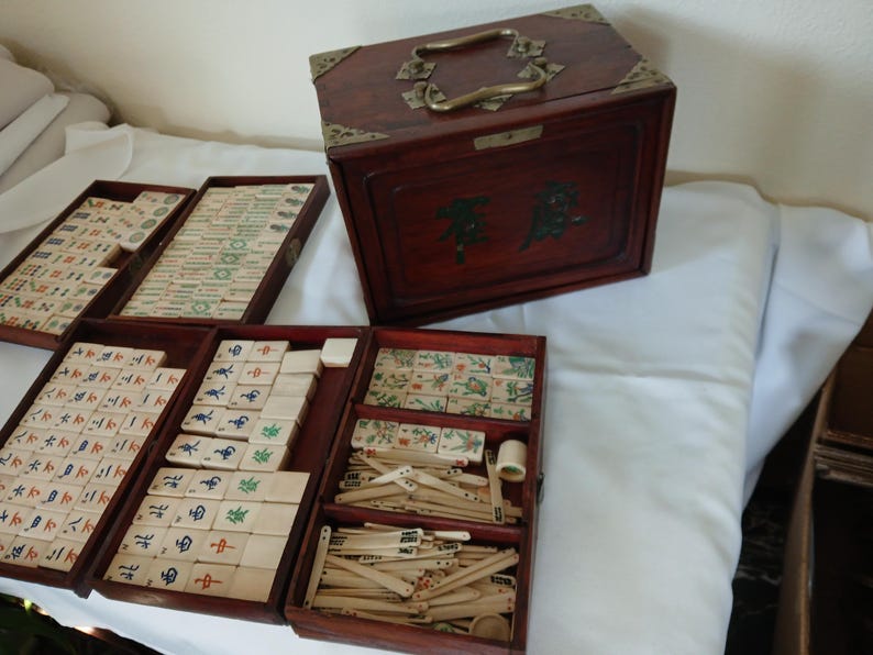 Puede incluir: Un juego de Mahjong de madera vintage con un acabado rojo oscuro. El juego incluye una caja de madera con herrajes de lat&oacute;n y varias bandejas llenas de fichas y accesorios. Las fichas presentan caracteres y s&iacute;mbolos chinos en varios colores.