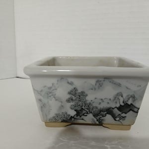 May include: A white ceramic square bonsai pot with a black and gray mountain scene design. The pot has four small feet.