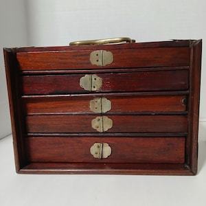 Puede incluir: Una caja de joyer&iacute;a de madera vintage con cuatro cajones. La caja es de un color marr&oacute;n oscuro intenso con herrajes de lat&oacute;n, incluidos tiradores de caj&oacute;n y un asa. Los cajones son rectangulares y tienen un dise&ntilde;o simple y cl&aacute;sico.