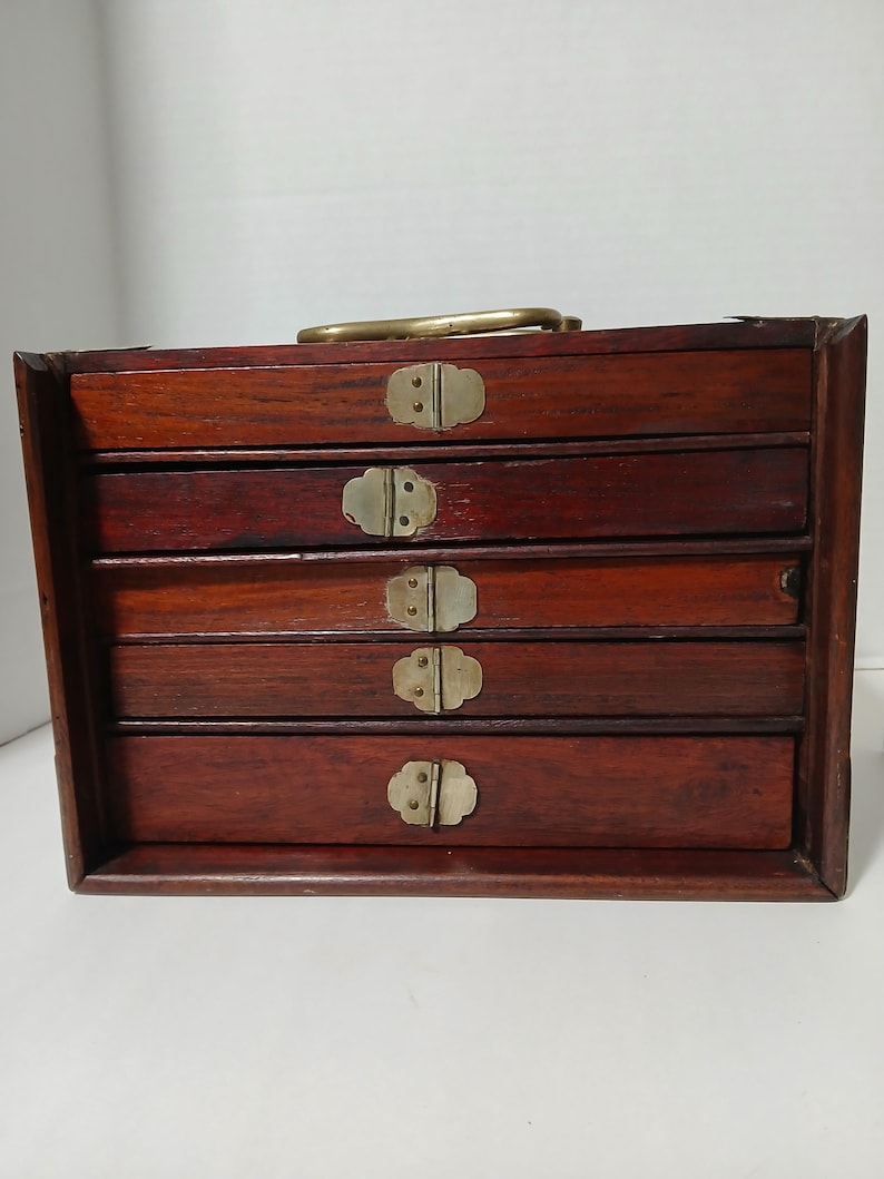 Puede incluir: Una caja de joyer&iacute;a de madera vintage con cuatro cajones. La caja es de un color marr&oacute;n oscuro intenso con herrajes de lat&oacute;n, incluidos tiradores de caj&oacute;n y un asa. Los cajones son rectangulares y tienen un dise&ntilde;o simple y cl&aacute;sico.