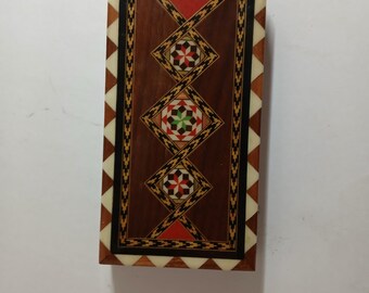 May include: Rectangular wooden box with a decorative inlaid design. The box features a dark brown central panel with three diamond-shaped patterns, each containing a star-like design in red, green, and black. The edges are bordered with white and brown triangles.