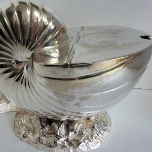 Puede incluir: Un bol de servir de plata con forma de concha de nautilus con tapa. El bol está sobre una base que se asemeja a un lecho de roca con algas marinas y peces.