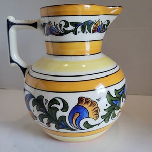 Może przedstawiać: Ceramiczny dzbanek w kolorze żółtym i białym z kwiatowym wzorem w kolorze niebieskim, zielonym i pomarańczowym. Dzbanek ma szeroki otwór i uchwyt.