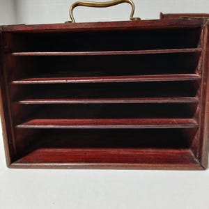Puede incluir: Un organizador de madera de color marr&oacute;n rojizo oscuro con m&uacute;ltiples ranuras horizontales. Cuenta con un asa de color lat&oacute;n en la parte superior. El organizador parece estar dise&ntilde;ado para guardar documentos o art&iacute;culos peque&ntilde;os, ofreciendo un dise&ntilde;o cl&aacute;sico y funcional.
